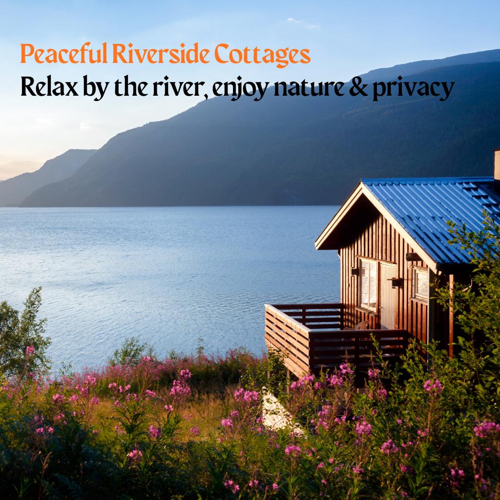 A close-up view of a small, rustic wooden cottage with a dark blue metal roof, nestled on a grassy bank near a wide, calm river or lake. The cottage has a small wooden deck and is surrounded by lush green foliage and patches of purple-pink wildflowers. In the background, the large body of water stretches out to the base of imposing, tree-covered mountains under a bright sky with the sun setting or rising. Text overlay reads: "Peaceful Riverside Cottages" in orange and "Relax by the river, enjoy nature & privacy" in black.